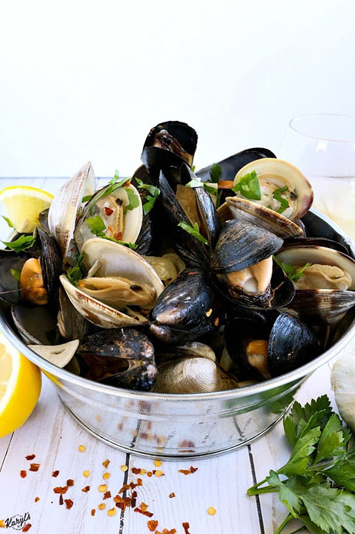 picture of clams and mussels in metal bucket, with lemons, garlic, parsley & red pepper flakes on table and glass of wine in backgriound