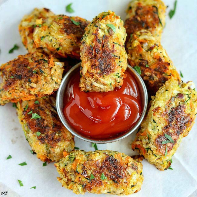 Baked Zucchini Carrot Tots on white paper, surrounding a metal container with ketchup