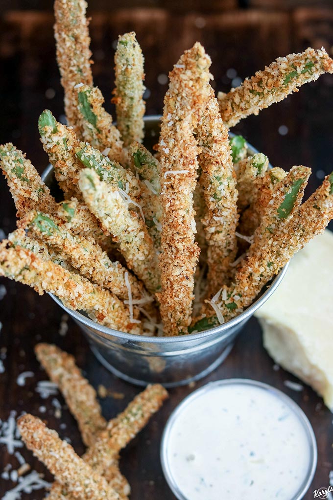 close up shot of Baked Green Bean Fries in a metal bucket with a side of ranch in front and a block of Parmesan cheese on the side
