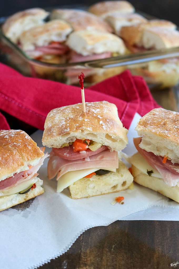 close up shot of Hot Muffaletta Sliders on white paper with baking dish of sliders in the background 