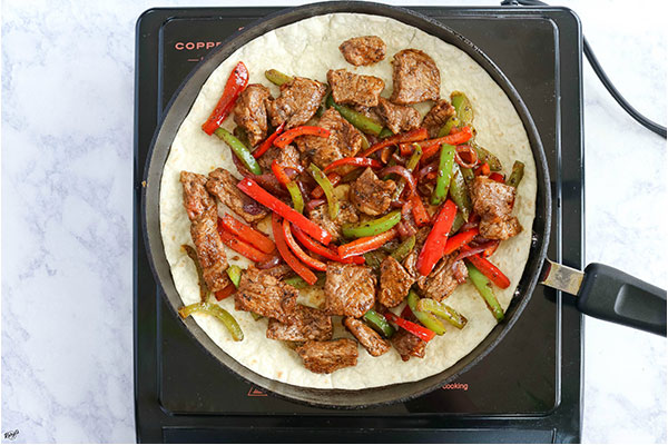 overhead process shot: tortilla in a black skillet, topped with steak, onion and pepper mixture
