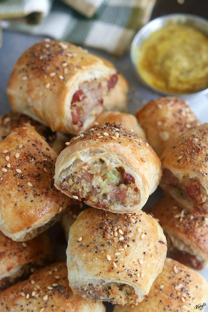 close up shot of finished sausage rolls in a pile on white paper. Side of mustard in a metal container in back right corner