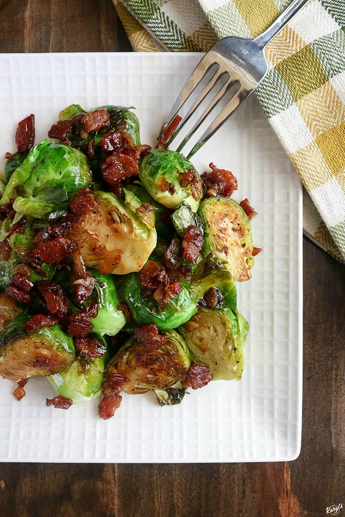overhead shot of finished Bourbon Bacon Brussel Sprouts on a white plate with a metal fork and a green/yellow/white napkin on the side