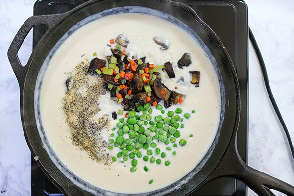 overhead process shot: peas, vegetables and seasonings added to cream sauce in a black skillet