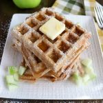 Angled shot of a stack of finished Apple Cinnamon Waffles on a white plate with 3 apples in the background