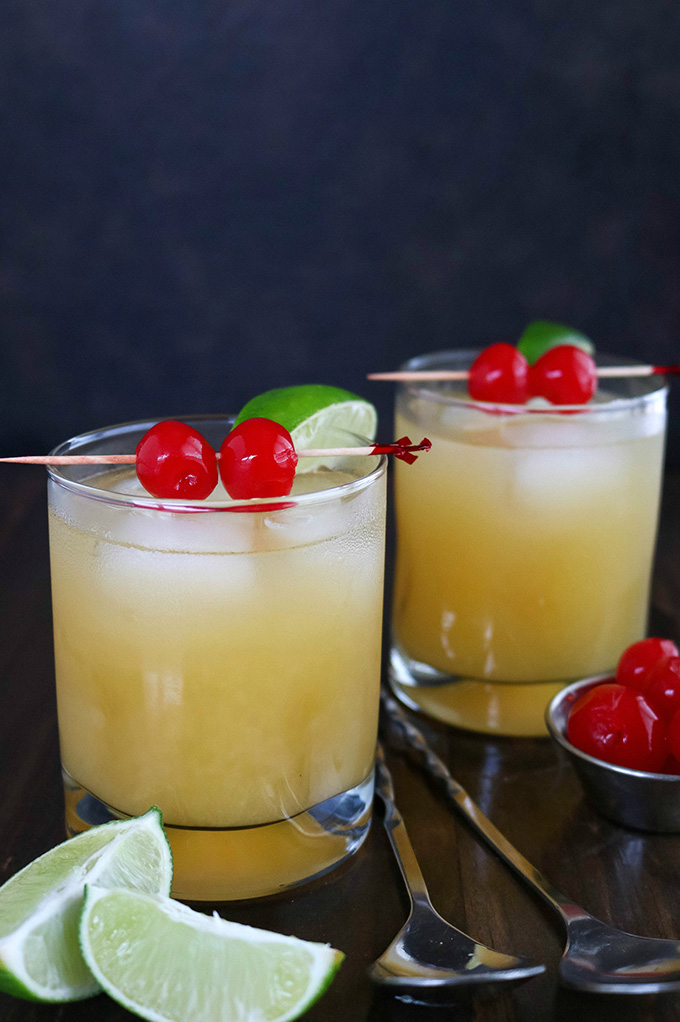 shot of 2 glasses of Black Eyed Susan Cocktails with lime wedges and cherries on the side